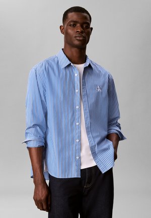 Man draagt een blauw-wit verticaal gestreept overhemd met knopen over een wit T-shirt en een donkere broek, staand tegen een effen grijze achtergrond.