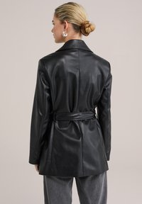 Svart läderblazer med midjebälte, långa ärmar och klassisk krage. Slät textur med elegant design och skräddarsydd passform.