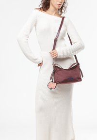 Bordeaux handtas met een gladde, matte afwerking, voorzien van een bloemaccessoire, schouderriem en een blauw merklogo. G Combineerd met een witte geribbelde jurk.