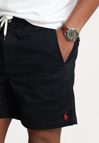Person som bär svarta shorts med röd Polo-logotyp och ett grönt armbandsur med brunt läderrem, hand i fickan.