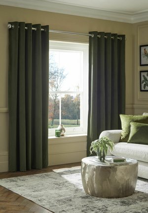 Wohnzimmer mit dunkelgrünen Vorhängen, weißem Fenster, hellem Sofa mit grünen Kissen, rundem metallischem Couchtisch und Topfpflanze auf Teppich.