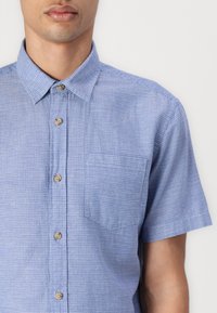 Chemise à manches courtes, avec boutons, de couleur bleu clair avec un motif texturé, dotée d'une poche poitrine et de boutons en bois.