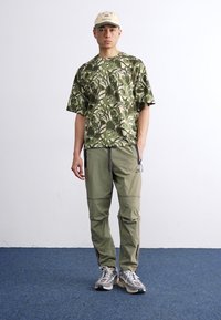 Camo-mønstret grøn t-shirt med korte ærmer, parret med lysegrønne bukser. Model iført en beige kasket og grå sneakers med accenter.