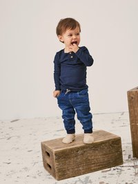 Marine langermet skjorte med to knapper, denimbukser med elastiske mansjetter, står på en treboks, iført beige strikkesokker.