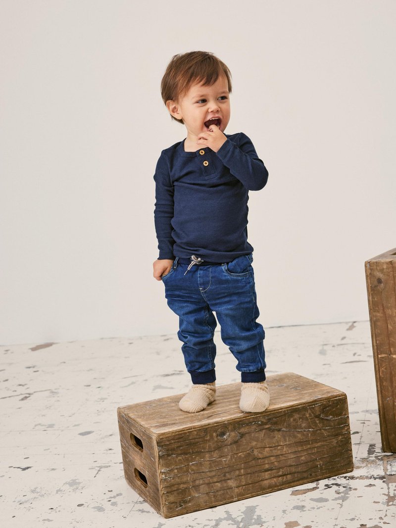 Marine langermet skjorte med to knapper, denimbukser med elastiske mansjetter, står på en treboks, iført beige strikkesokker.