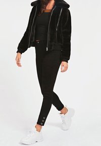 Giacca corta in ecopelliccia nera con chiusura a zip, abbinata a jeans neri aderenti con dettagli a bottone alla caviglia e sneakers bianche.