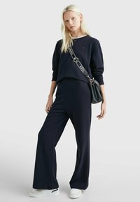 Felpa navy e pantaloni palazzo abbinati in morbido tessuto, con colletto rotondo bianco e logo minimal. Accostati a una borsa nera piccola.