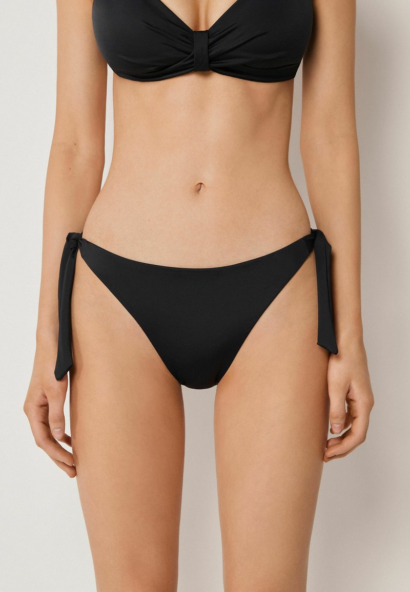Mujer con bikini negro con lazada lateral en la parte inferior y top bandeau fruncido, de pie contra un fondo liso y claro.