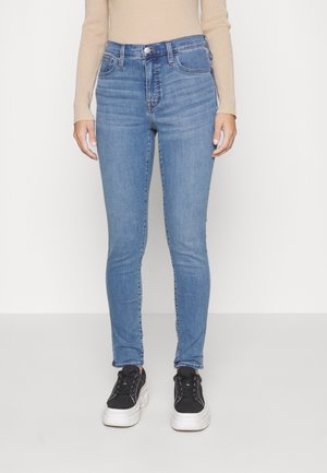 Model iført lyseblå skinny jeans med falmede detaljer, sorte platform sneakers og en beige ribbet langærmet top mod en ensfarvet baggrund.