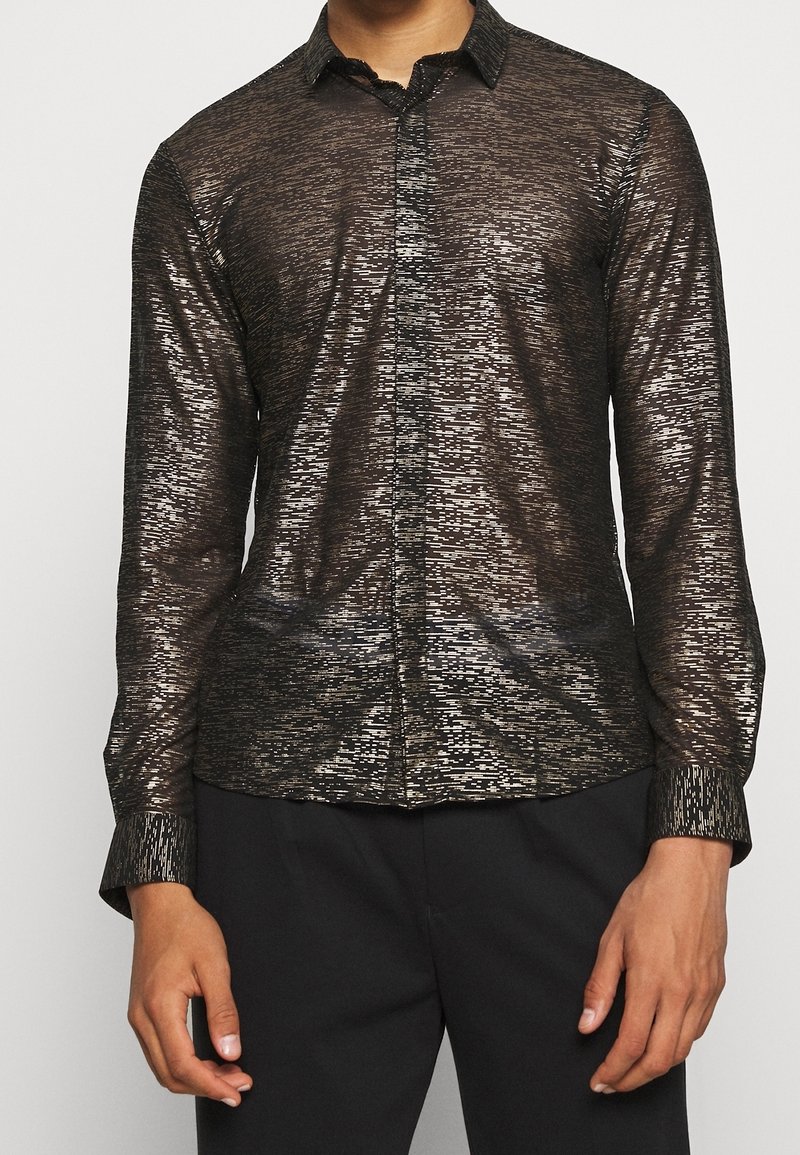 Homme portant une chemise noire à manches longues avec un motif métallique et boutons, pantalon noir, mains relâchées le long du corps.