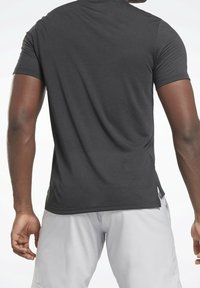 Camiseta deportiva negra de manga corta con textura lisa, dobladillo curvado y aberturas laterales para mayor flexibilidad, combinada con pantalones cortos de color claro.