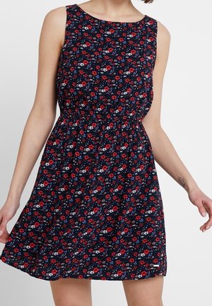 Vestido azul marino sin mangas con estampado floral en rojo, blanco y azul, cintura ceñida, llevado por una persona de pie visible desde los hombros hasta la mitad del muslo.