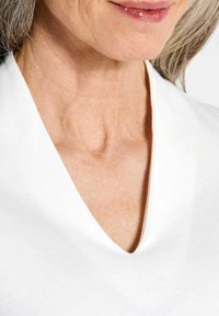 Witte V-hals top gemaakt van zachte stof, met een gladde textuur en strakke lijnen rond de halslijn, die een subtiele draping onthult.