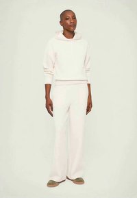 Person med barbert hode iført en hvit hettegenser, matchende vide bukser og olivengrønne sko, stående foran en ensfarget lys bakgrunn.