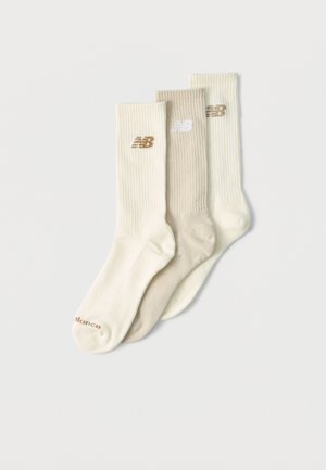 Tre paia di calzini a coste a girocollo in crema, beige e bianco sporco con il logo New Balance sui polsini disposti sovrapposti su uno sfondo bianco.