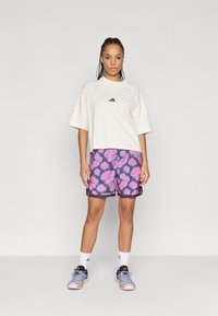 Λευκό oversized μπλουζάκι με μαύρο λογότυπο στο στήθος, συνδυασμένο με μωβ σορτς που φέρουν ροζ σχέδια καρδιάς. Το μοντέλο φοράει γκρι αθλητικά παπούτσια και λευκές κάλτσες.