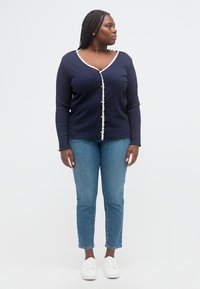 Γυναίκα με πλεξούδες, φοράει μια μπλε σκούρα ζακέτα με λευκό περίγραμμα, μπλε τζιν και λευκά αθλητικά παπούτσια, στέκεται και κοιτάζει προς τα πλάγια σε ένα απλό φόντο.