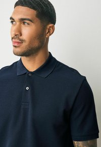 Marinen polo shirt lavet af tekstureret stof, med en klassisk krave, to knapper og korte ærmer. Subtil branding detalje synlig.