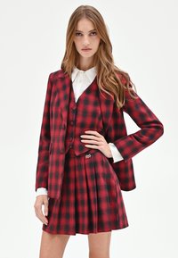 Completo a quadri rosso e nero con blazer aderente, gilet abbottonato e minigonna plissettata. Presenta polsini di camicia bianchi e un dettaglio con logo sulla gonna.