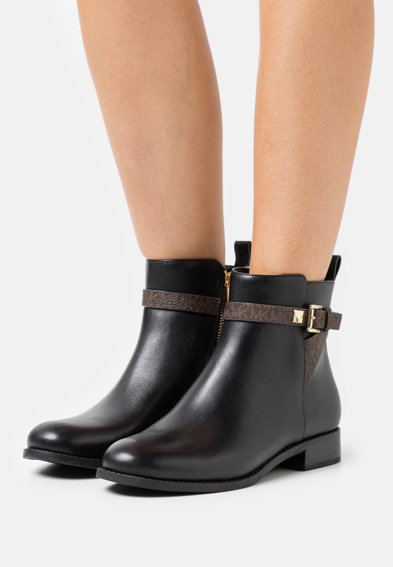 Bottes montantes en cuir noir avec des sangles texturées marron et des boucles dorées portées sur des jambes nues, avec des fermetures éclair latérales et des talons bas.