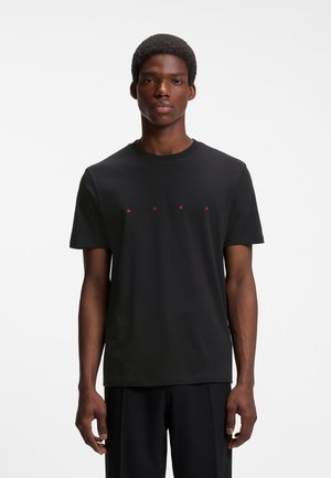 Junger Mann trägt ein einfaches schwarzes T-Shirt mit roten Buchstaben, die HUGO über der Brust schreiben, und steht vor weißem Hintergrund.