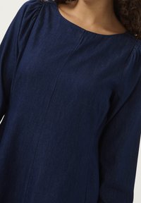 Mörkblå denimtop med långa ärmar, båtformad halsringning och subtila rynkdetaljer vid axlarna. Slät textur och rak siluett.