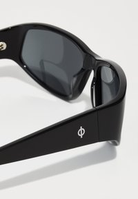 Occhiali da sole neri con lenti scure e finitura lucida. Presentano un distintivo simbolo sulla parte laterale. Design elegante e moderno con una forma curva.