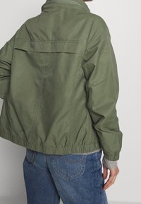 Olivengrøn jakke med løs pasform, elastisk kant, lynlåslukning foran og lagdelt lommendesign. Lavet af let, struktureret stof.
