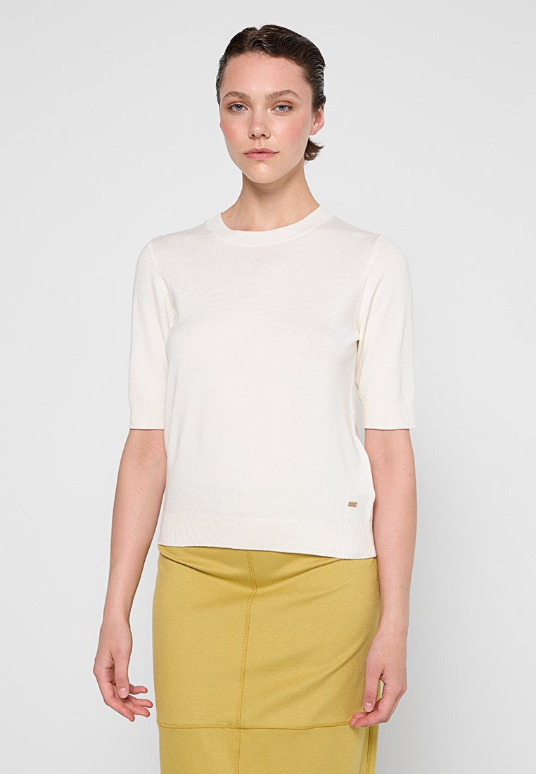 Pull blanc à manches courtes avec un col rond, poignets et ourlet côtelés, associé à une jupe jaune. Présente un petit logo en accent à l'ourlet.