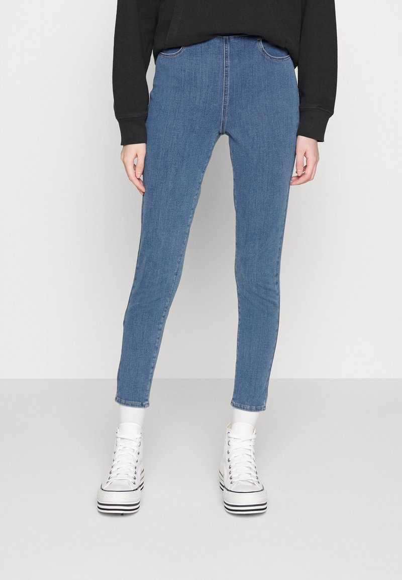 Skinny blue denim jeans with a mid-rise waist, featuring five-pocket styling and subtle stitching details, paired with white high-top sneakers.