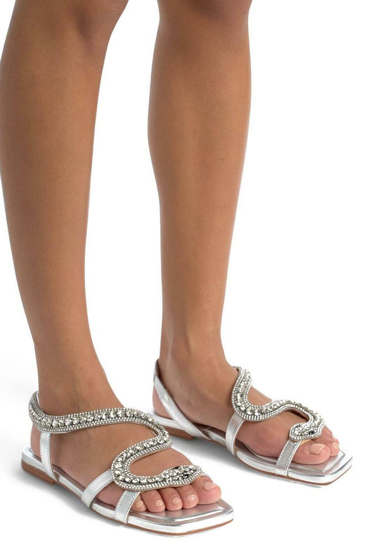 Le sandali piatti argento presentano una cinghia ondulata adornata con strass. Il design include una punta aperta e una finitura liscia e lucida.