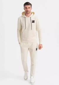 Beige Kapuzenpullover mit einem schwarzen Logo-Patch, Kordeln und einer Fronttasche. Kombiniert mit passenden beigen Jogginghosen, die einen Logo-Patch und einen elastischen Bund haben.