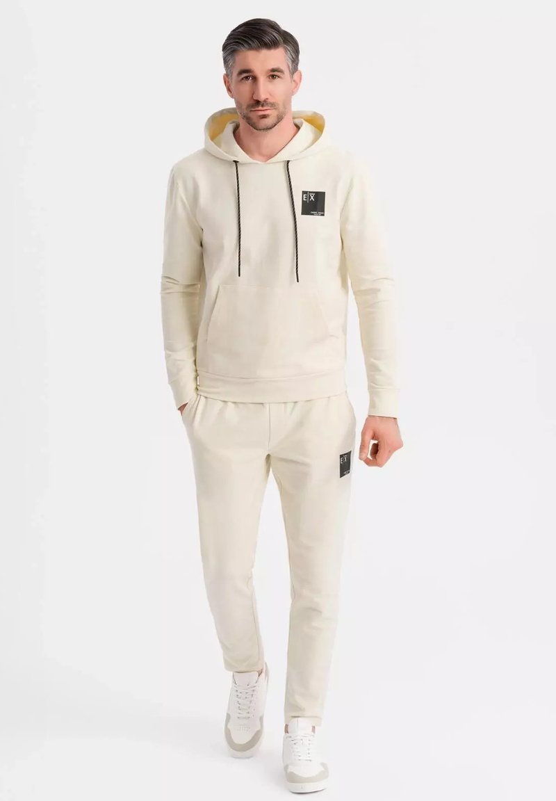 Beige Kapuzenpullover mit einem schwarzen Logo-Patch, Kordeln und einer Fronttasche. Kombiniert mit passenden beigen Jogginghosen, die einen Logo-Patch und einen elastischen Bund haben.