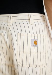 Gestreifte Canvas-Shorts in Creme mit navyfarbenen Linien, ausgestattet mit einer Gesäßtasche und einem Carhartt-Logo-Patch. Robustes Material, lässiges Design.