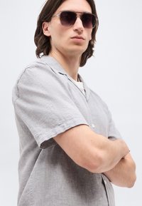 Νέος άνδρας με μακριά σκούρα μαλλιά μέχρι τους ώμους, φοράει σκούρα γυαλιά ηλίου και ανοιχτό γκρι πουκάμισο με κοντά μανίκια και κουμπιά, με σταυρωμένα τα χέρια.