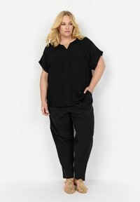 Haut noir à manches courtes avec un col en V et une couture centrale, associé à un pantalon noir ample. Les chaussures sont de couleur claire avec des détails découpés.