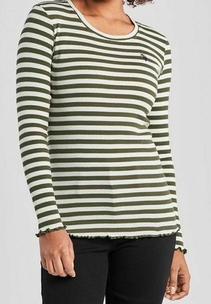 Femme portant un haut ajusté à manches longues rayé olive et blanc avec des bords en forme de laitue aux poignets et à l'ourlet, assorti à un pantalon noir.