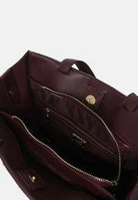 Sac à main bourgogne avec un extérieur en cuir lisse, des compartiments zippés et des accessoires en métal doré. L'intérieur comprend plusieurs poches et une doublure en satin.