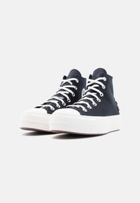 Ζευγάρι μαύρα ψηλά πάνινα παπούτσια με λευκές λαστιχένιες σόλες και λευκά κορδόνια, εμφανισμένα με κλίση σε λευκό φόντο.