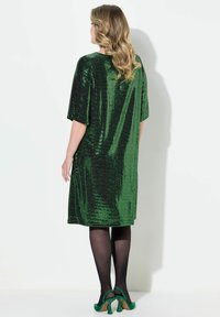 Grünes Pailletten-Kleid mit kurzen Ärmeln, lockerer Passform, schulterlang und glänzender Textur; kombiniert mit schwarzen Strumpfhosen und grünen Absätzen.