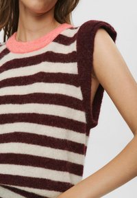 Top fără mâneci din tricot moale, cu dungi orizontale în nuanțe de vișiniu și crem, având un decolteu moale, contrastant, de culoare roz pal, purtat de o persoană cu pielea deschisă.