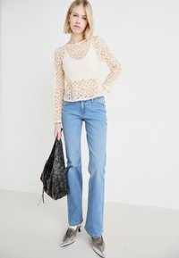 Top de encaje de mangas largas con patrones circulares, combinado con vaqueros acampanados de color azul claro. Lleva un bolso negro texturizado y calza tacones metálicos.