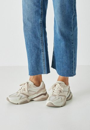 Klobige Sneakers in hellem Beige mit Mesh- und Wildleder-Akzenten, ausgestattet mit einer dicken Sohle und Schnürsenkeln, kombiniert mit gekürzten blauen Jeans.