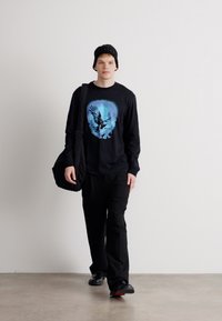 Cămașă neagră cu mânecă lungă și un design grafic al unui păsări de culoare albastră, asortată cu pantaloni negri și o geantă de umăr. Modelul poartă o caciulă neagră.