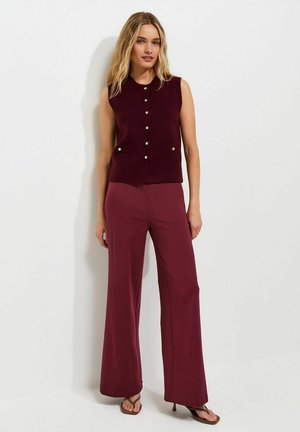 Vrouw met blond haar die een mouwloos, bordeauxkleurig, button-down topje draagt en bijpassende wijde broek met sandalen met hak, staand tegen een witte muur.