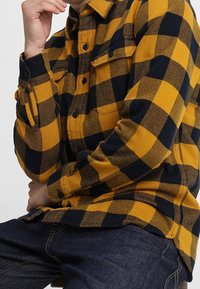 Camicia di flanella a scacchi gialli e neri con bottoni sul davanti, due tasche sul petto e un aspetto morbido e textured.