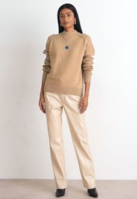 Suéter bege de malha com textura canelada e patch de logótipo, combinado com calças de couro brancas e sapatos pretos de bico fino.