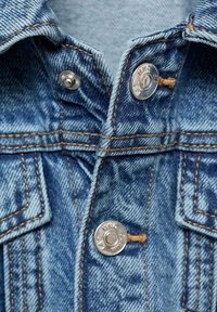 Pohled zblízka na přední stranu modré džínsové bundy, která ukazuje stříbrné kovové knoflíky s vyrytým textem a oranžové švy.