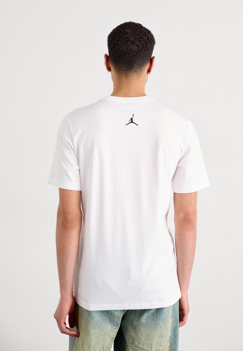 T-shirt blanc à manches courtes avec un logo Jumpman noir dans le haut du dos. Le tissu semble doux avec une coupe standard et un design à col rond.