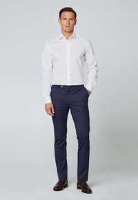 Hvid langærmet skjorte med knapper, kombineret med mørkeblå skræddersyede bukser og brune sko. Minimalistisk design og klassisk pasform.